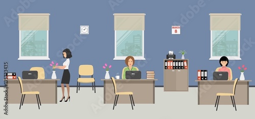 Office room on the day of March 8. The young women are employees at work. There is beige furniture on a window background in the picture. There are vases with flowers on the tables also. 