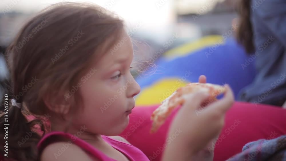Beautiful cute little girl sitting in bean bag chairs and eating pizza at the park in summer day. Side view. Beautiful cute little girl sitting in bean bag chairs and eating pizza at the park in summer day. Side view.
