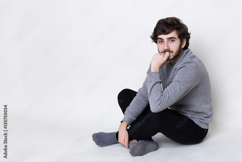 A portrait of handsome man having dark beard with moustache wearing casual clothes sitting on the floor crossed legs holding his hand on his chin looking into camera with mysterious look A portrait of handsome man having dark beard with moustache wearing casual clothes sitting on the floor crossed legs holding his hand on his chin looking into camera with mysterious look