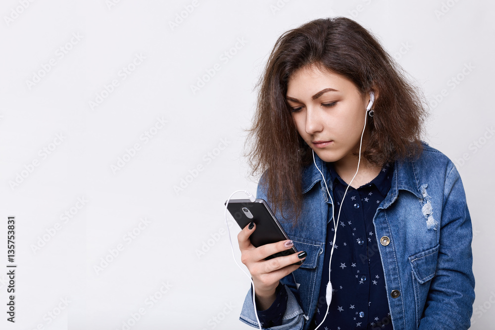 People, technology and teens concept. A pretty young girl with her smartphone and earphones listening to the music looking attentively in her cell-phone having a rest and enjoing music People, technology and teens concept. A pretty young girl with her smartphone and earphones listening to the music looking attentively in her cell-phone having a rest and enjoing music