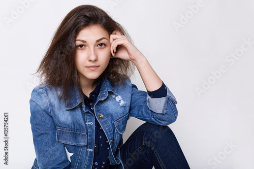 Obraz na plátne A close-up of attractive young brown-eyed girl wearing jean jacket looking into camera very attentively being confident in herself holding  hand on her head