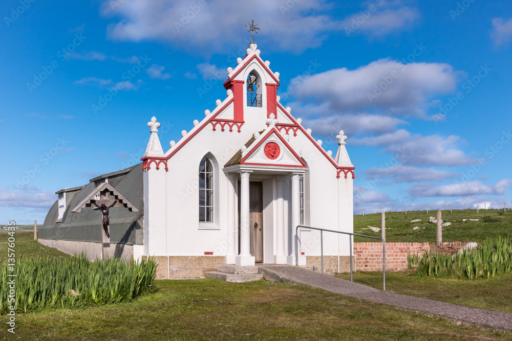 Orkneys, Scotland - June 5, 2012: The entire building, entrance and front facade of the white and maroon painted Italian Chapel on Lamb Holm Island. Green lawn in front, against deep blue sky.