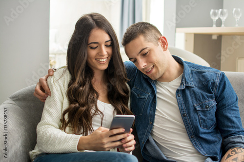 Happy young couple looking at smartphone together while sitting on couch at home.