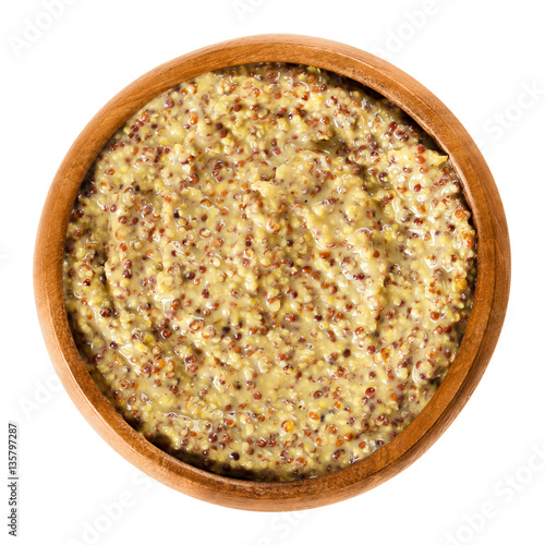 Whole grain Dijon mustard in wooden bowl with brown seeds. Traditional mustard named after the town of Dijon in Burgundy, France. Isolated macro food photo close up from above on white background.