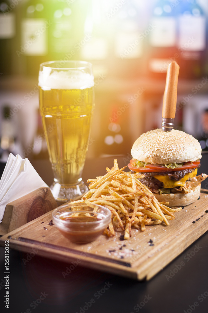 Tasty grilled cheeseburger with a knife stabbed trough on a wooden plate. Napkins and beer on the side. Lens flare in the background. Tasty grilled cheeseburger with a knife stabbed trough on a wooden plate. Napkins and beer on the side. Lens flare in the background.