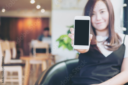 Mockup image of a beautiful woman holding and showing white mobile phone with blank black screen and smiley face in modern cafe