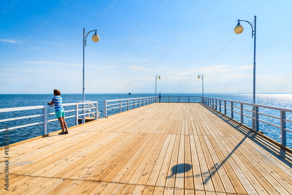 Wooden pier and young woman standing against railing in distance in Jurata town on coast of Baltic Sea, Poland