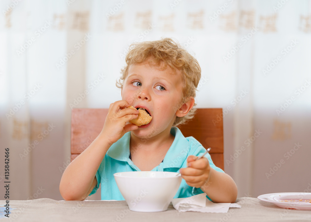 Blonde boy having breakfast while sitting on a chair at the kitchen table at home. White porcelain cup in front of kid. Bites off a piece of white bread. Holds spoon with his hand. Blonde boy having breakfast while sitting on a chair at the kitchen table at home. White porcelain cup in front of kid. Bites off a piece of white bread. Holds spoon with his hand.