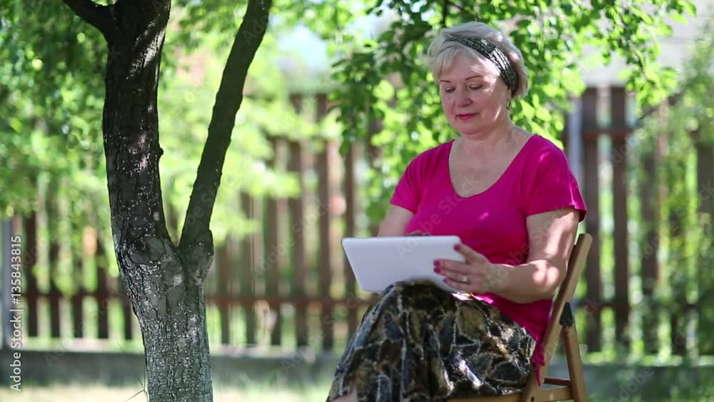 Blonde woman sits on the chair in garden and uses tablet computer. Senior woman with tablet pc sits under tree in garden. Female with tablet computer Blonde woman sits on the chair in garden and uses tablet computer. Senior woman with tablet pc sits under tree in garden. Female with tablet computer