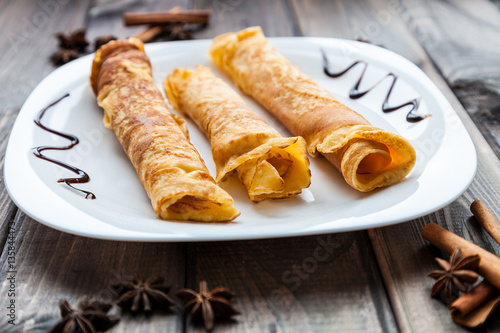 Sweet fresh homemade crepes with chocolate topping on dark grey rustic wooden table. Menu for breakfast, desserts and lunch. Cinnamon and kitchen herb for decoration.
