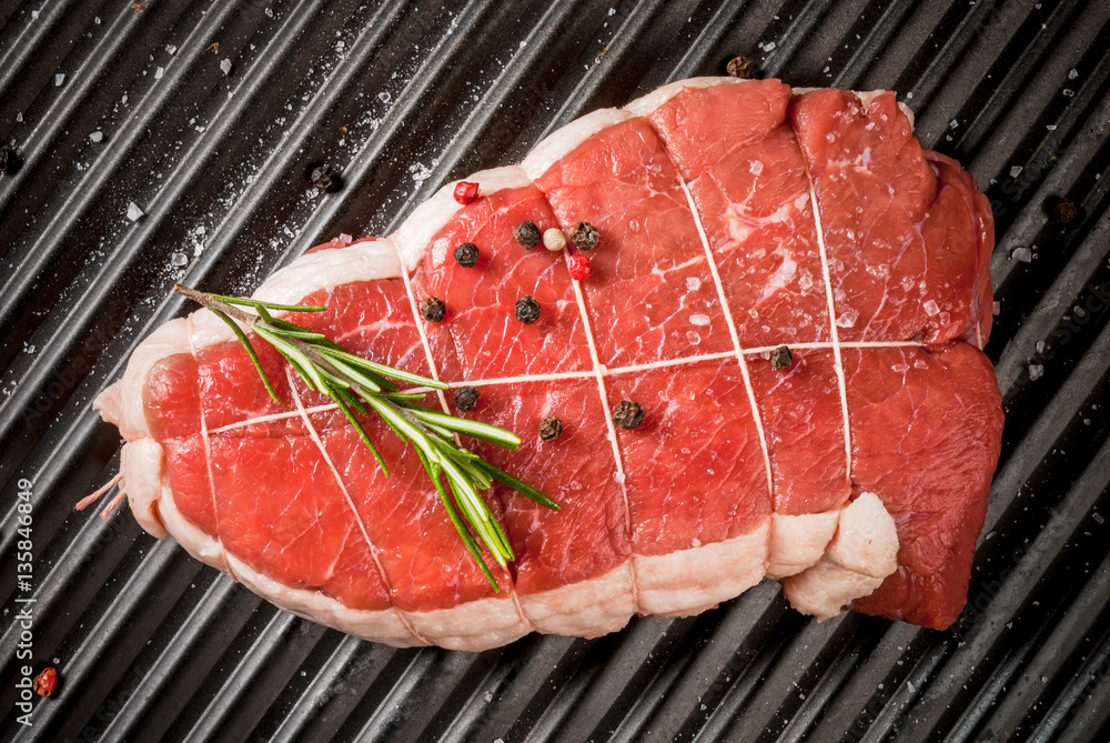 Fresh uncooked roast beef on on grill pan with salt, pepper and rosemary for cooking. Top view Fresh uncooked roast beef on on grill pan with salt, pepper and rosemary for cooking. Top view