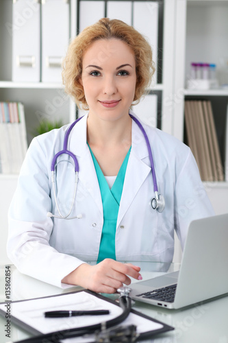 Happy blonde female doctor sitting at the table and typing by laptop computer. Medicine, healthcare and help concept. Physician ready to examine patient