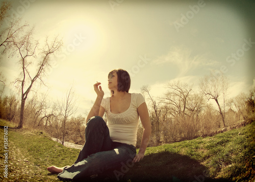 White girl with brown hair sits next to a river and trees smoking a cigarette. Girl wearing white t-shirt and jeans, barefoot sitting on the grass. Fish eye lens and instagram filter effect added.