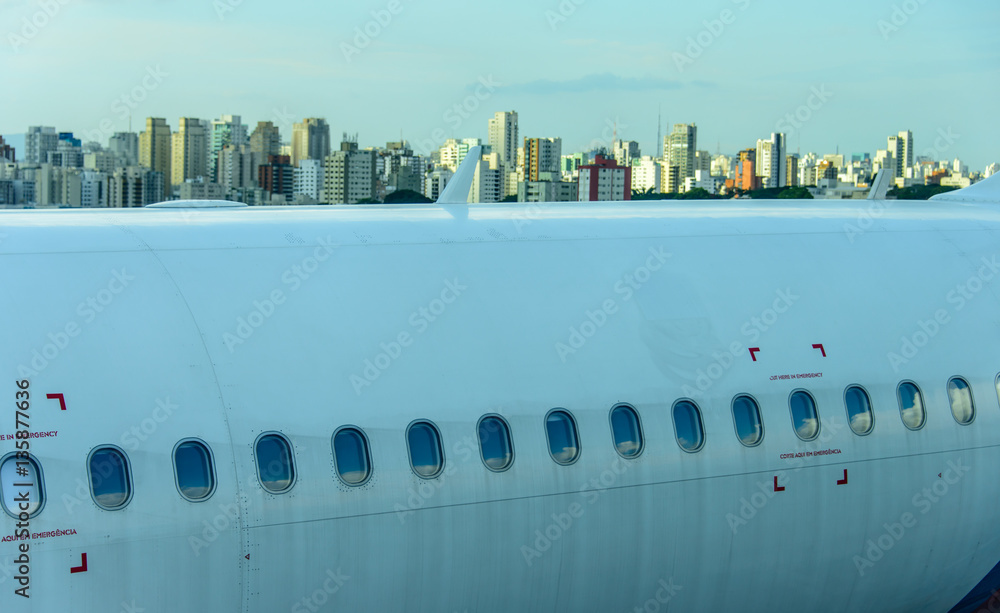 Fuselage of the narrow-body aircraft with white clouds and blue sky reflected in the portholes at the airport on the background of high-rise modern buildings Fuselage of the narrow-body aircraft with white clouds and blue sky reflected in the portholes at the airport on the background of high-rise modern buildings