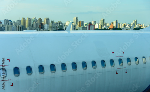 Fuselage of the narrow-body aircraft with white clouds and blue sky reflected in the portholes at the airport on the background of high-rise modern buildings