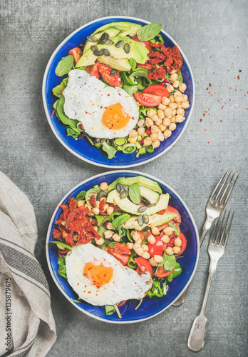 Healthy breakfast with fried egg, chickpea sprouts, seeds, fresh vegetables and greens in blue bowls over grey concrete background, top view. Clean eating, healthy lifestyle, vegetarian food concept