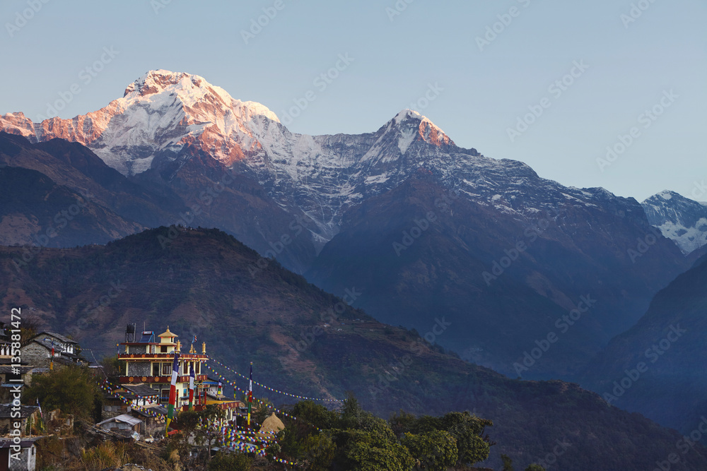Rural life in front of gigantic Himalayas, Nepal. Incredible view of Nepali village with Buddhist flags strung along, spectacular ancient mountains with white craggy peaks standing high in background