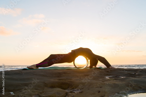 Girl in red bottoms, gray jacket and black hat doing yoga on a green yoga mat near the ocean at sunset with yoga wheel