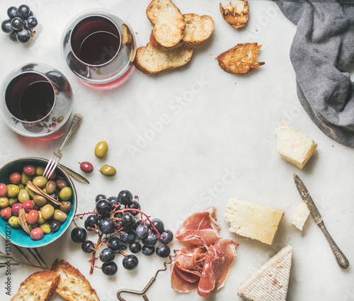 Wine and snack set. Variety of cheese, olives in blue ceramic bowl, prosciutto meat, roasted baguette slices, black grapes and glasses of red wine over grey marble background, top view, copy space
