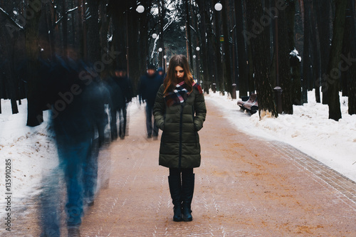 Portrait of a beautiful young girl in a jacket, a scarf tied around the neck, frozen in time, in the middle of the park alley on a background distorted by passing people.