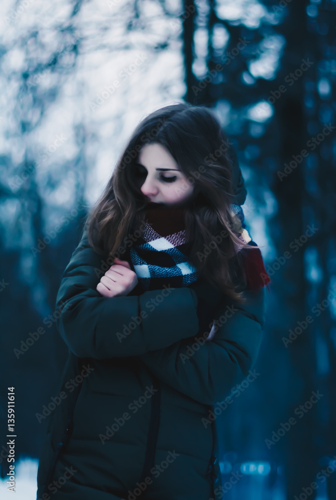 Portrait of a beautiful brown-haired young girl, with pale face, red lips, in a jacket and a scarf tied around the neck, on the background of dark trees. Portrait of a beautiful girl. Portrait of a beautiful brown-haired young girl, with pale face, red lips, in a jacket and a scarf tied around the neck, on the background of dark trees. Portrait of a beautiful girl.