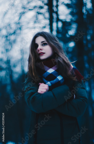 Portrait of a beautiful brown-haired young girl, with pale face, red lips, in a jacket and a scarf tied around the neck, on the background of dark trees. Portrait of a beautiful girl.
