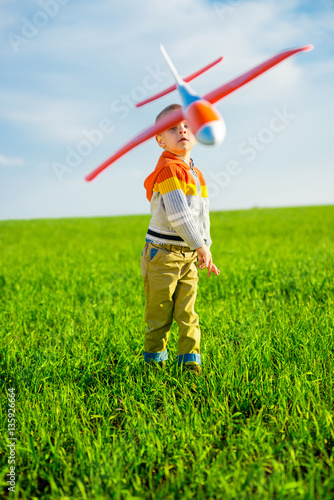 Happy kid playing with toy airplane against blue summer sky background. Boy throw foam plane in green field. Best childhood concept.