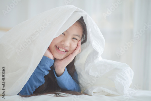 Happy smiling Asian girl playing hide and seek under the blanket in her bed room
