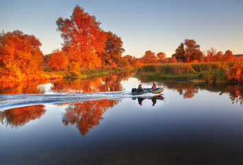 Fishermen sailing fast at rubber powerboat by river making trace at tranquil water surface against beautiful morning scenery with golden color foliage trees at bank. Fishing background.