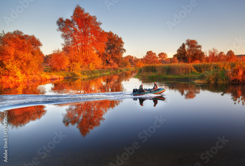 Fishermen sailing fast at rubber powerboat by river making trace at tranquil water surface against beautiful morning scenery with golden color foliage trees at bank. Fishing background.