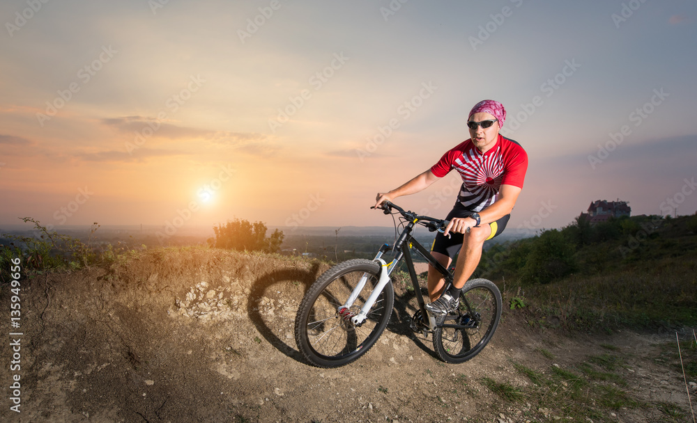 Man in glasses riding on the mountain bicycle on a trail against evening sky with bright sun on the sunset