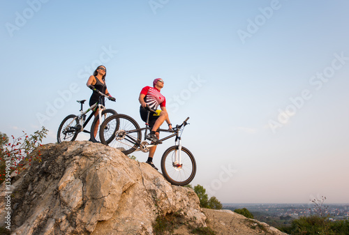 Young couple bikers in sunglasses staying on the bicycles at the precipice of hill against evening sky and looking at the sunset. Bottom view