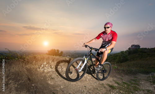 Fototapeta Naklejka Na Ścianę i Meble -  Man in glasses riding on the mountain bicycle on a trail against evening sky with bright sun on the sunset