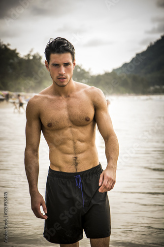 Half body shot of a handsome young man standing on a beach in Phuket Island, Thailand, shirtless wearing boxer shorts, showing muscular fit body