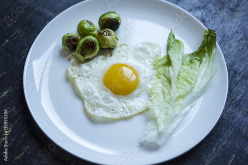 fried egg with cucumbers, olives, and brussels sprouts on a plate with fork and knife on a dark wooden background