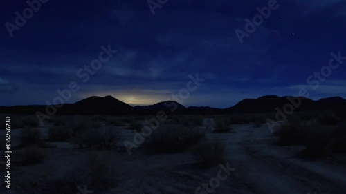 Wide angle time lapse night in Mojave desert along dirt road with clouds and planes passing in sky with city lights glowing in distance