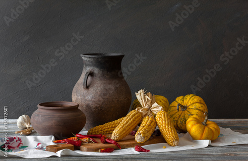 Still life in a rustic style: autumn harvest: set of clay dishes, corn, pumpkins and wooden spoon on a table. Natural light from a window.