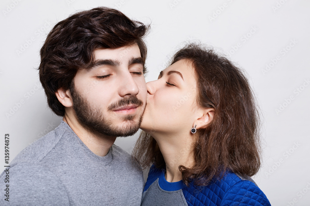 Romantic loving couple. A pretty brunette kissing his bearded boyfriend in his chin. A happy couple closing their eyes from pleasure isolated over white background having romantic evening Romantic loving couple. A pretty brunette kissing his bearded boyfriend in his chin. A happy couple closing their eyes from pleasure isolated over white background having romantic evening