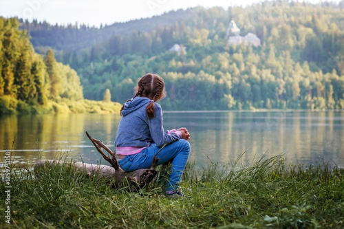 A girl child sits on the shore of a mountain lake.