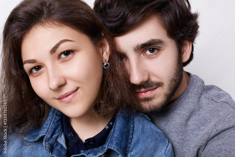 A close-up of two young people in love over white background. A bearded hipster embracing her girlfriend with charming eyes and smile. Happy to be together. Fashionable couple hugging