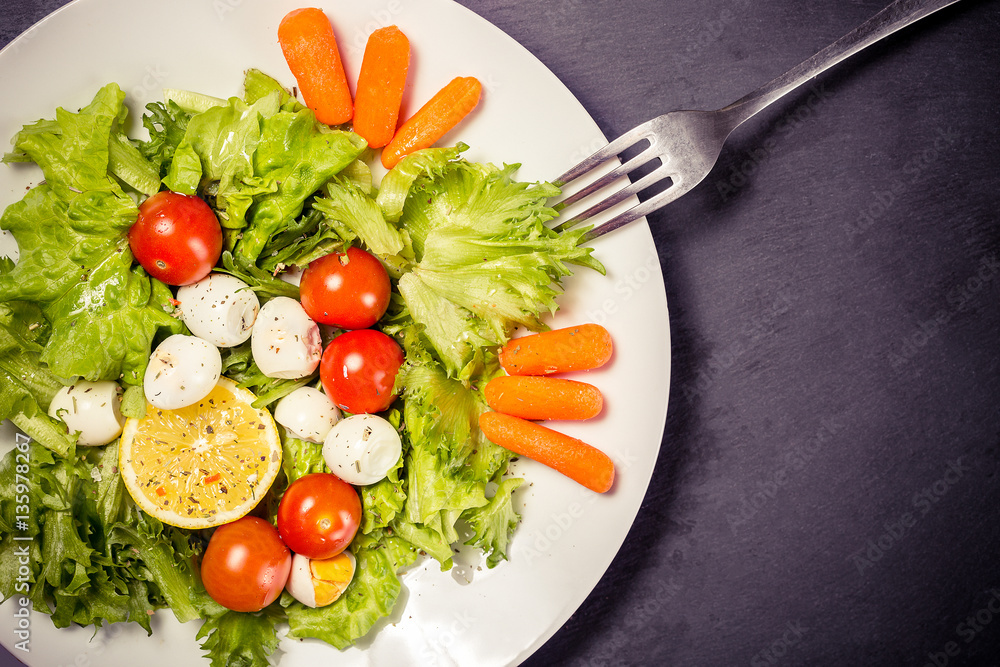Tasty salad with cherry tomatoes, salad leaves, lemon, spices, carrot and quail eggs on stone table with fork. Top view Tasty salad with cherry tomatoes, salad leaves, lemon, spices, carrot and quail eggs on stone table with fork. Top view