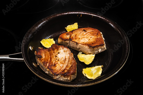 Two tuna steaks and lemon pieces in a frying pan on a black stove