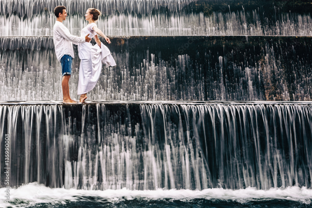 Fototapeta premium Happy family on honeymoon holidays - married loving couple hugging, kissing with fun under falling water in cascade waterfall pool. Active lifestyle, people travel on summer vacation on Bali island.