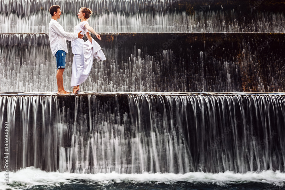 Fototapeta premium Happy family on honeymoon holidays - married loving couple hugging, kissing with fun under falling water in cascade waterfall pool. Active lifestyle, people travel on summer vacation on Bali island.