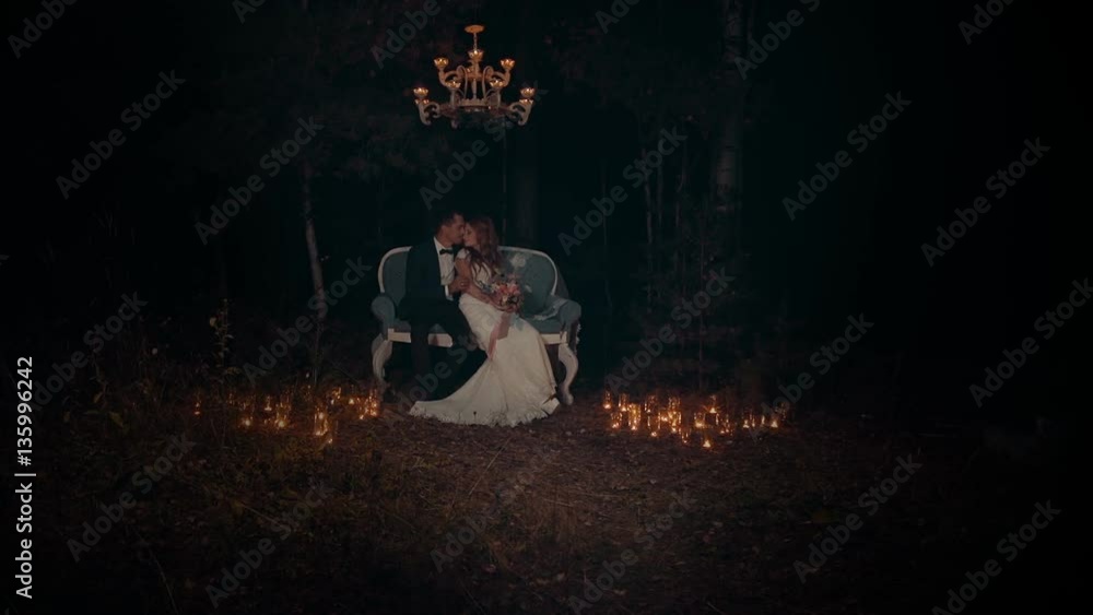 The newlyweds embrace and kiss at night, sitting on a luxurious couch under a white chandelier with lights in a dark forest illuminated by bright lit candles in the grass. The newlyweds embrace and kiss at night, sitting on a luxurious couch under a white chandelier with lights in a dark forest illuminated by bright lit candles in the grass.