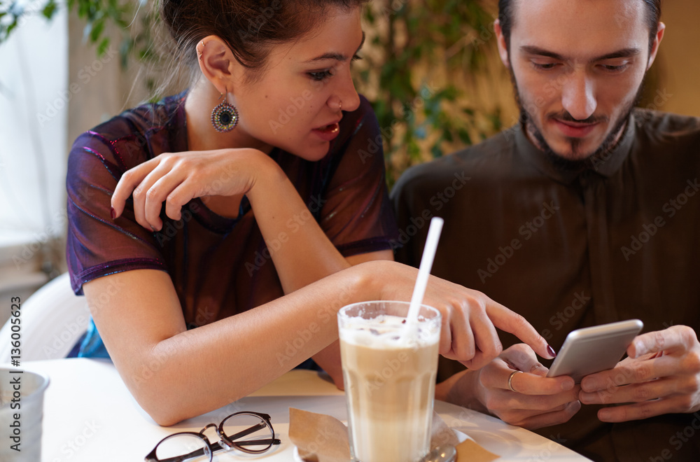 Attractive young friends sitting in a cafe looking at a smartphone. Hipster man show something to his girlfriend on the phone. Beautiful male and female person discuss the news from social networks Attractive young friends sitting in a cafe looking at a smartphone. Hipster man show something to his girlfriend on the phone. Beautiful male and female person discuss the news from social networks
