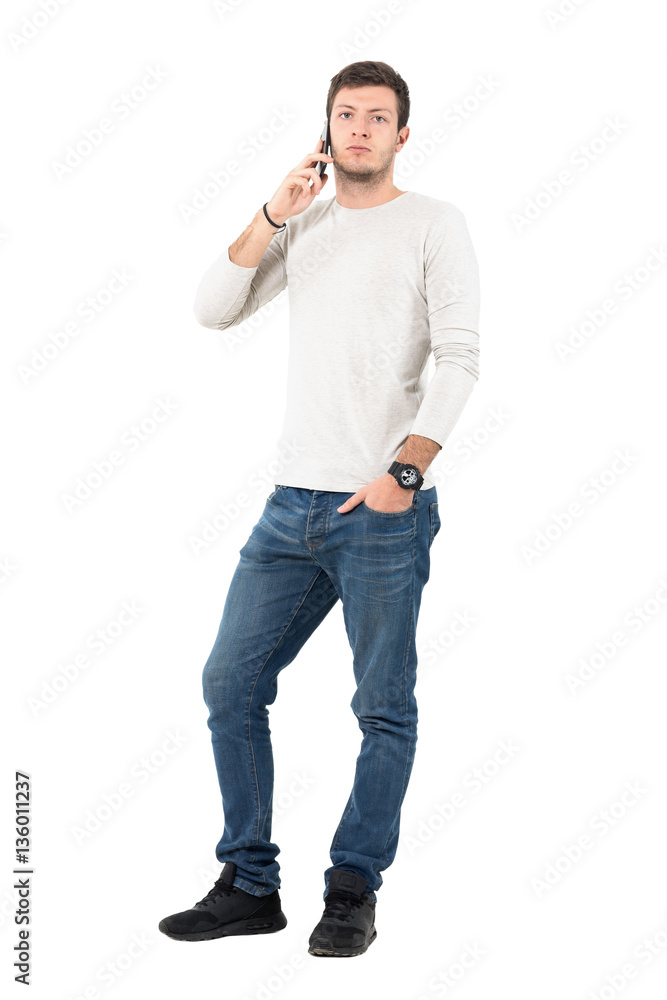 Cool young man talking on the mobile phone looking at camera with hand in pocket. Full body length portrait isolated over white studio background. Cool young man talking on the mobile phone looking at camera with hand in pocket. Full body length portrait isolated over white studio background.