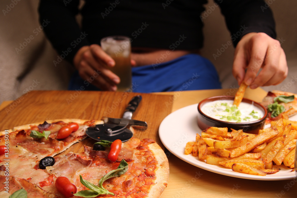 Fat man sitting on a sofa eating fries at the table with pizza, candy, sandwiches and chips. is unhealthy lifestyle.  junk food.
