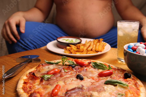 Fat man sitting on a sofa at the table with pizza, candy, sandwiches and chips. is unhealthy lifestyle.  junk food.
