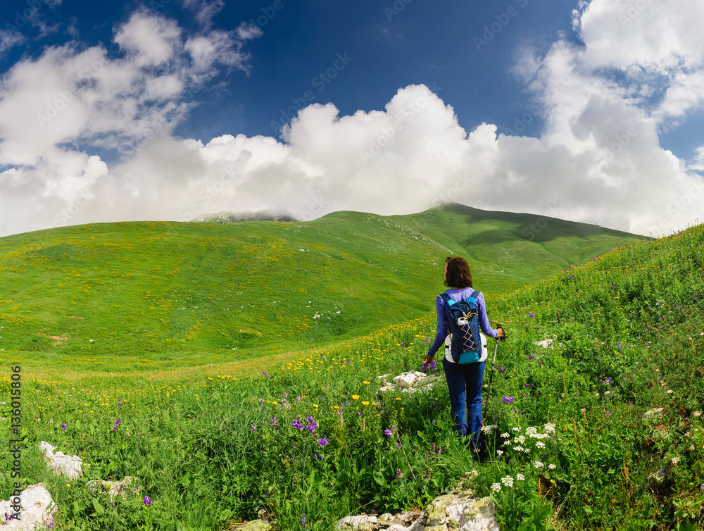 Tourist girl walking on a greenery hill trail with a backpack and hiking poles
